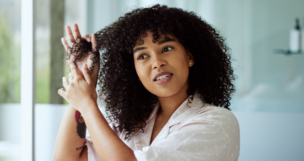 Mulher com cabelo cacheado castanho escuro mexendo no seu cabelo.